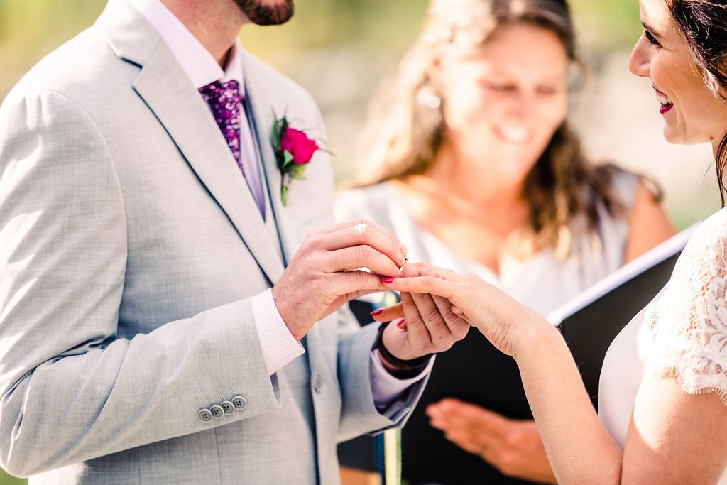 Sarah Harding smiling in background as groom puts wedding ring on brides finger